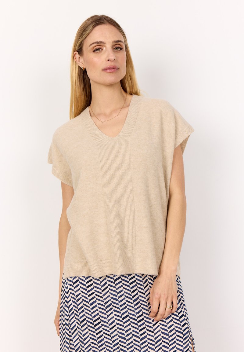 Beige stickad tröja med V-ringning, korta ärmar och ribbade detaljer. Modellen bär den över en mönstrad kjol med blå och vita zigzag-mönster.