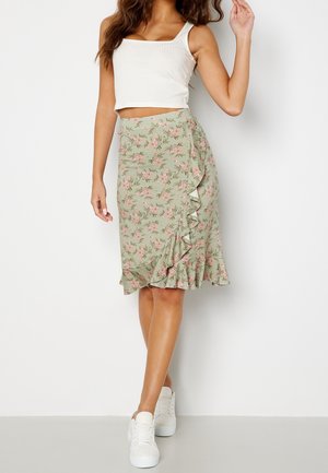 Mujer con top blanco sin mangas, falda verde floral con volantes y zapatillas blancas, de pie frente a fondo blanco.