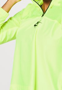 Veste légère vert néon avec zip sur le devant, texture en maille et logo Nike noir sur la poitrine. Comprend une capuche et des poignets élastiques.