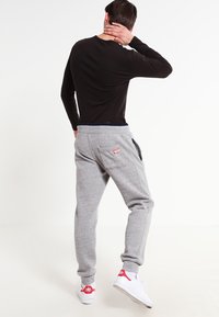Schwarzes langärmeliges Hemd und graue Sweatpants mit umgeschlagenen Bündchen am Knöchel. Die Sweatpants haben seitliche Taschen und einen roten Logo-Patch auf der Rückseite. Weiße Turnschuhe.