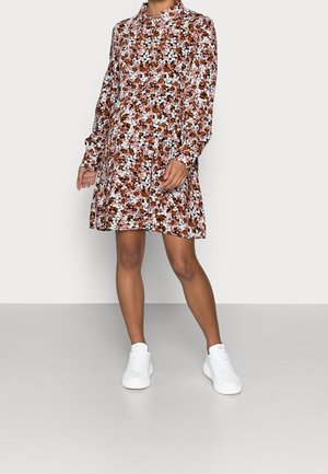 Blumenkleid mit langen Ärmeln in einer Mischung aus Rosa, Braun und Weiß. Es hat einen Kragen und endet oberhalb des Knies, kombiniert mit weißen Sneakers.