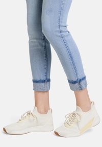 Vita atletiska sneakers med en texturerad stickad ovandel, snörning och gult accent på sidan, tillsammans med ljusblå jeans som är uppvikta och har dekorerade muddar.