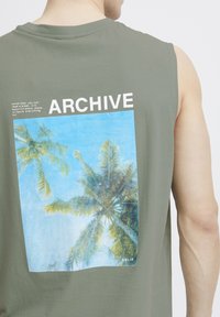 Ærmeløs olivengrøn skjorte med et rygmotiv af palmer mod en blå himmel, med ordet "ARCHIVE" trykt ovenfor.