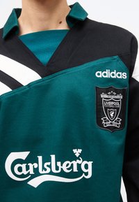 Gros plan sur un maillot de football du Liverpool FC de couleur bleu sarcelle et noir, avec le logo Adidas et le sponsor Carlsberg sur la poitrine.