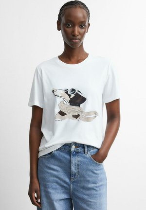 Frau trägt ein weißes T-Shirt mit einer Grafik eines Hundes mit Fliegerbrille und Schal, kombiniert mit blauen Jeans, steht vor einfachem Hintergrund.