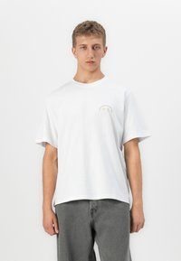 Ung mann med kort blondt hår iført en hvit T-skjorte med en liten logo på brystet og vide grå jeans, stående mot hvit bakgrunn.
