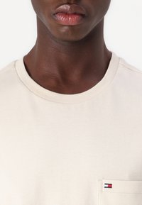 Beige cotton T-shirt with a round neckline and a pocket featuring a small red, white, and blue logo on the pocket. Smooth texture.