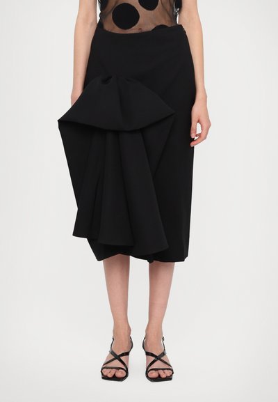 Falda negra con un diseño plisado escultórico en la parte frontal, hecha de un material texturizado. Llevada con sandalias negras de tiras.