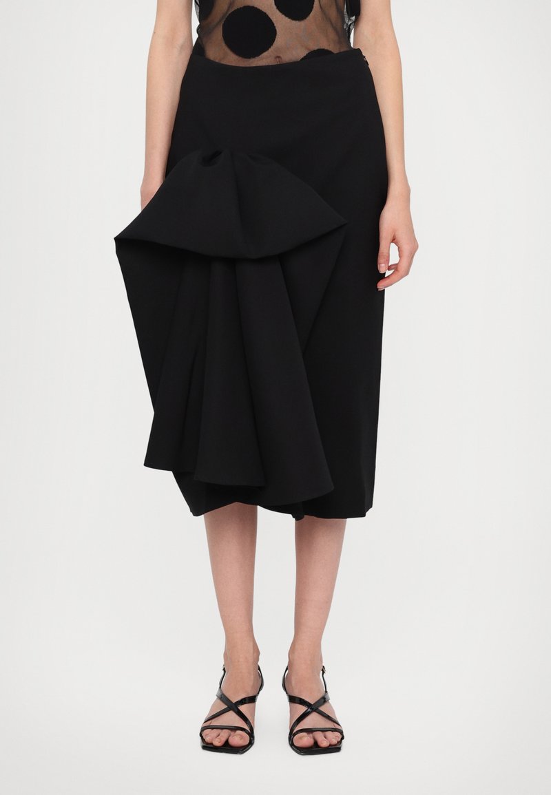 Black skirt with a sculptural pleated design at the front, made of textured material. Worn with black strappy sandals.