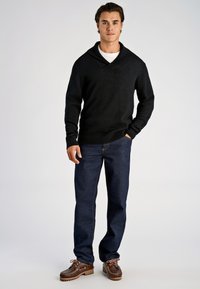 Schwarzer Pullover mit Schalkragen und gerippten Bündchen, kombiniert mit dunkelblauer Jeans und braunen Schnürschuhen. Glatte Stoffstruktur und figurbetontes Design.