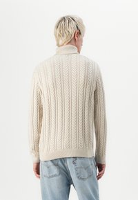 Maglione a trecce di colore crema, con colletto alto e polsini a costine, realizzato in tessuto a maglia testurizzata. Indossato con jeans blu chiaro.