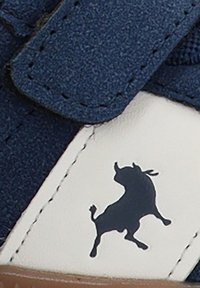 Zapatilla de color azul marino con un cierre de velcro, que presenta una sección blanca con un logotipo de toro negro y una superficie texturizada. Suela de goma.