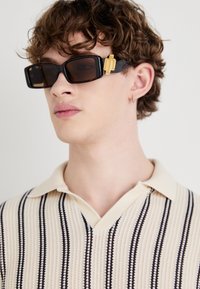 Occhiali da sole neri rettangolari con lenti ambra e accenti dorati sulle stanghette; indossati con una maglietta a polo in maglia a righe beige e blu navy.
