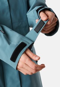 Giacca impermeabile blu con polsini regolabili dotati di cinturini in velcro neri. Texture liscia, vestibilità sagomata e design minimalista.
