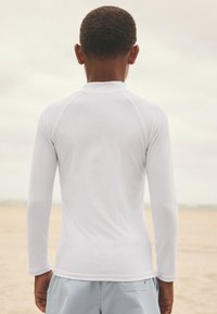 Långärmad, vit rashguard i mjukt, stretchigt tyg. Har hög krage och figursydd design med plana sömmar överallt.