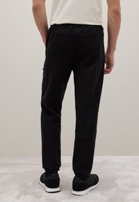 Schwarze Jogginghosen mit einem elastischen Bund, schmalem Schnitt, seitlichem Cargo-Taschen und strukturiertem Stoff, kombiniert mit schwarzen Sneakern auf einem hellen Hintergrund.