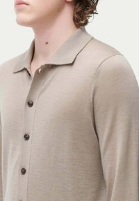 Beige stickad pikétröja med krage, med en knappslå och mörkbruna knappar. Slät textur och figurnära design.