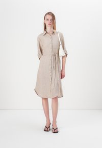 Robe chemise en lin beige avec un devant boutonné, des manches retroussées et une ceinture à la taille. Design minimaliste complété par un petit sac à main blanc.