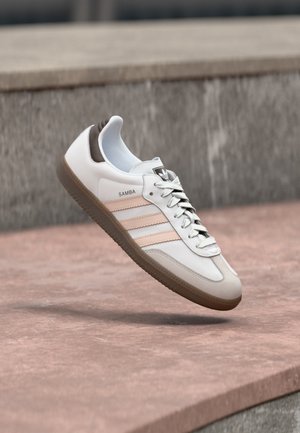 Baskets Adidas Samba blanches et beiges avec semelle en gomme flottant au-dessus d'une surface en pierre rosée et d'un fond en pierre grise.