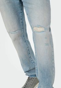Jean en denim bleu clair avec des détails déchirés, y compris des zones déchirées et effilochées au niveau des genoux, associés à des baskets grises.