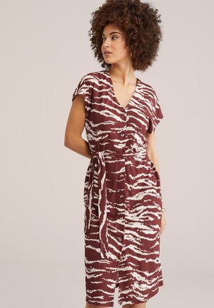 Femeie cu păr creț, purtând o rochie cu mâneci scurte, cu nasturi și curea, având un model abstract în nuanțe de roșu închis și alb.