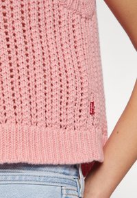 Pull en tricot rose avec une maille lâche et ouverte et un ourlet côtelé. Il présente une petite étiquette rouge de marque sur le côté. Stylé avec un jean bleu clair.