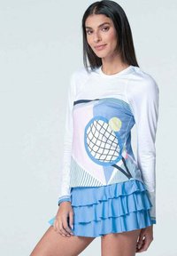 Frau in einem weißen Langarmoberteil mit einem Druck von Tennisschläger und Ball, kombiniert mit einem hellblauen, gestuften Rüschenshirt.