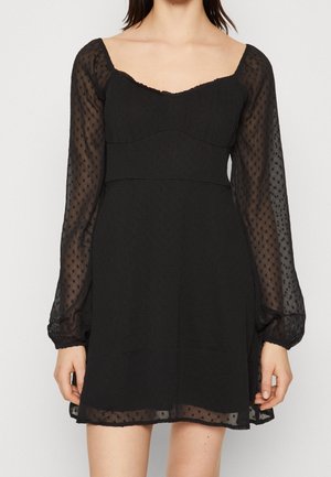 Kobieta w czarnej mini sukience z przezroczystymi długimi rękawami i dekoltem w kształcie serca, z subtelną wzorzystą fakturą w kropki.