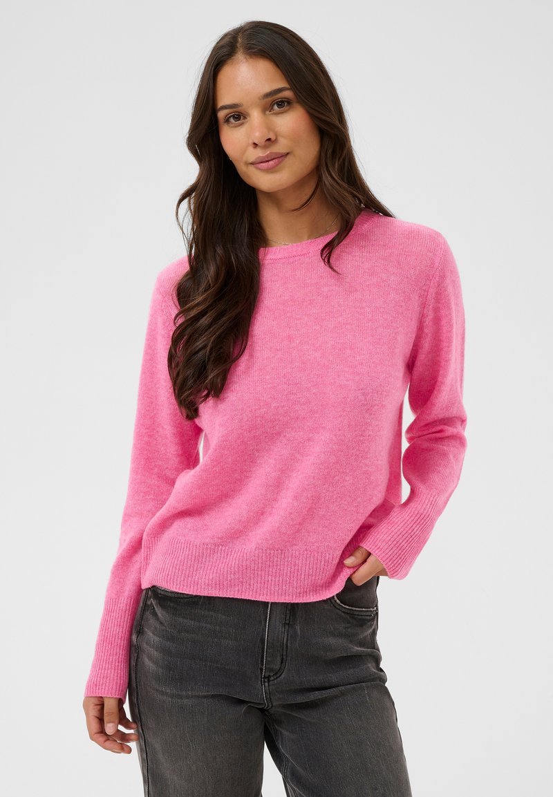 Pull en tricot rose avec un col rond, des poignets et un ourlet côtelés. Porté avec un jean noir, mettant en valeur une texture douce et une coupe décontractée.
