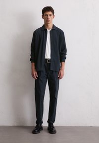 Camicia nera a bottoni sopra una maglietta bianca, abbinata a pantaloni scuri e scarpe nere. Design semplice con una vestibilità rilassata e minimi dettagli.