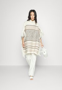 Poncho de malha em off-white com padrão geométrico bege, com franjas e um colarinho alto, combinado com calças largas brancas e uma bolsa pequena.