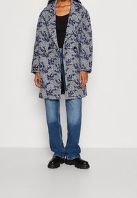 Vrouw in een blauw-wit patroon jas, zwarte top, blauwe jeans en zwarte chunky schoenen, staand tegen een effen achtergrond.