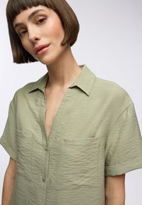 Camicia verde chiaro a maniche corte con tessuto testurizzato, chiusura frontale con bottoni e due tasche sul petto. Vestibilità rilassata e design con colletto classico.