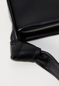 Črna usnjena torbica z gladko teksturo, opremljena s zvito naramnico in zadrgo vzdolž ene robu.