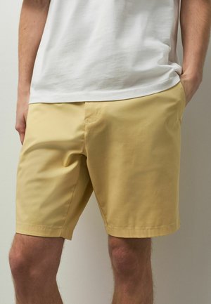 Mann trägt hellgelbe Shorts und ein weißes T-Shirt, steht mit einer Hand in der Tasche vor einem einfarbigen hellen Hintergrund.