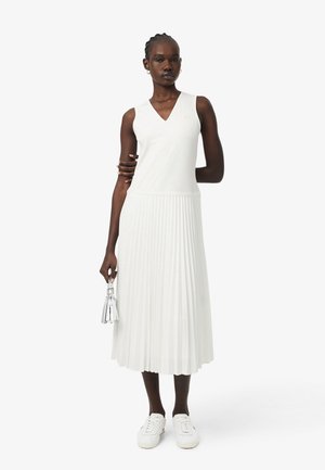 Frau trägt ärmelloses weißes V-Ausschnitt-Top und weißen plissierten Rock, hält silbernes Quasten-Accessoire, trägt weiße Turnschuhe, steht vor weißem Hintergrund.