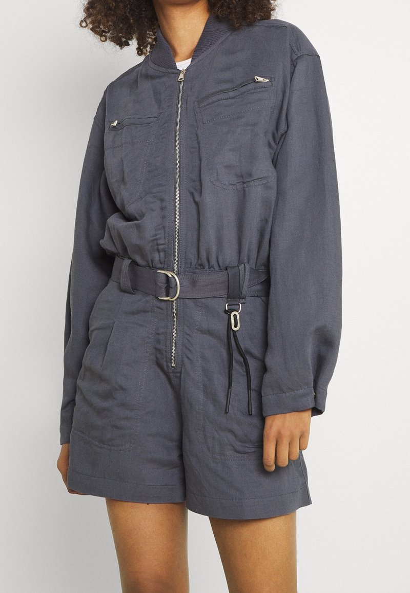 Femme portant une combinaison-short ceinturée gris foncé à manches longues, avec fermeture éclair devant, poches poitrine zippées et shorts amples.