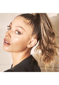 Närbild av en person med långt, vågigt brunt hår i en hög hästsvans, klädd i en svart topp och ett guldörhänge, med naturlig sminkning och gröna ögon.