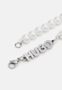 Gyöngyökkel díszített karkötő ezüst hardware-rel, az "HUGO" szó fényes fém betűkkel kiemelve, és homár csattal a zárásért.