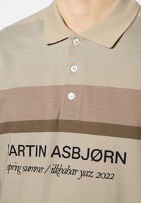 Polo en teintes beige sourdes avec des rayures horizontales en taupe et olive. Présente un col classique et une patte de boutonnage à trois boutons. Texte imprimé sur le devant.