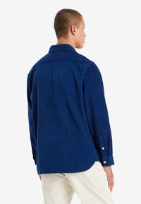 Camisa de pana azul marino de manga larga, con puños de botón y dobladillo redondeado, que presenta un cuello clásico y una textura suave.