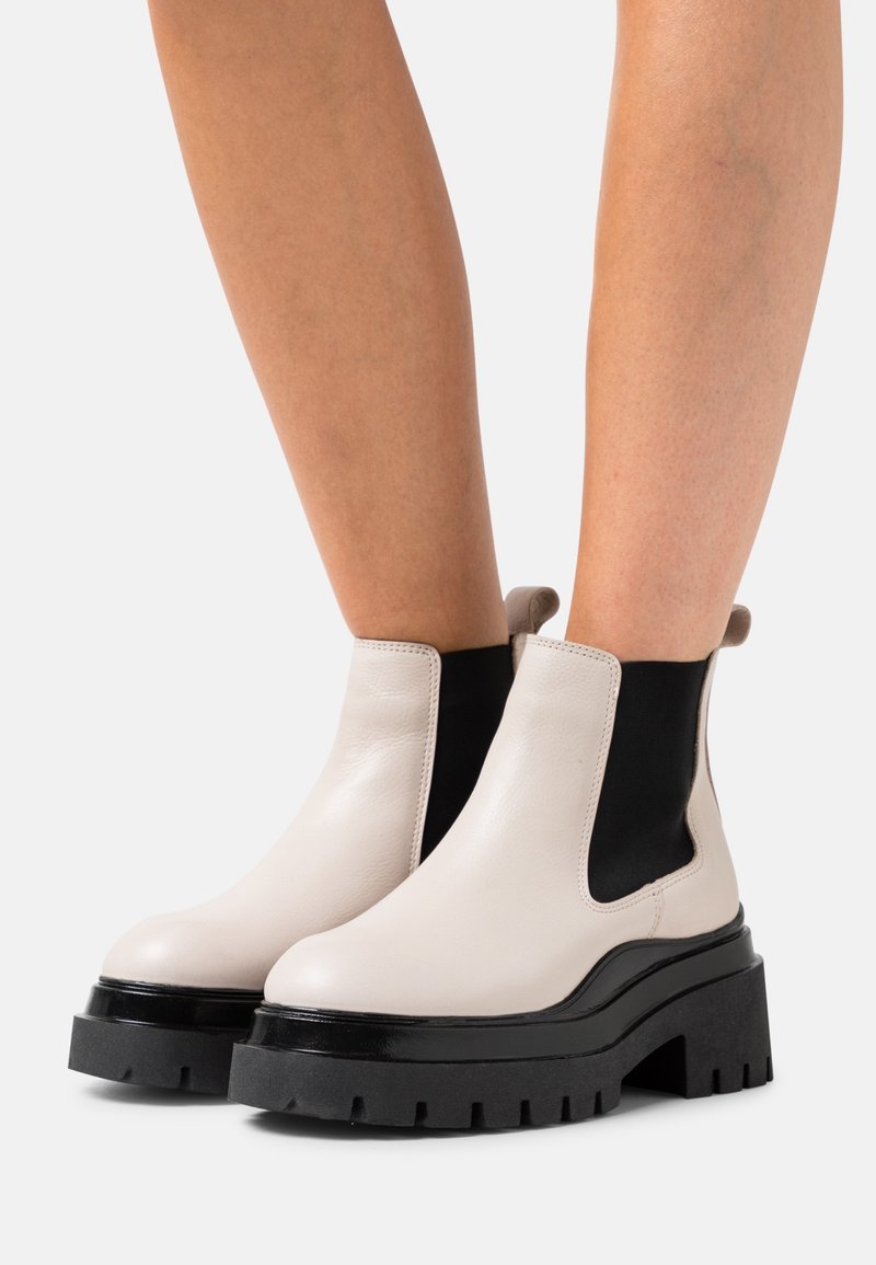 Bottes blanches en cuir montantes jusqu'à la cheville avec des semelles plateformes épaisses noires portées sur des jambes nues sur un fond uni.