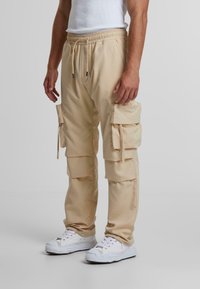 Beige cargo nadrág, elasztikus derekú, több oldalsó zsebbel, kényelmes fazonban. Fehér sportcipővel viselve. Sima textúra.