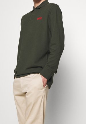 Polo à manches longues vert olive avec un patch de logo rouge, doté d'un col classique et d'une fermeture à boutons. Associé à un pantalon beige.