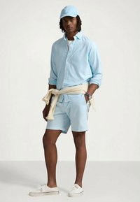 Homem com boné azul claro, camisa de manga longa abotoada, calções combinando, ténis brancos, com um suéter creme atado à cintura, em pé contra um fundo simples.