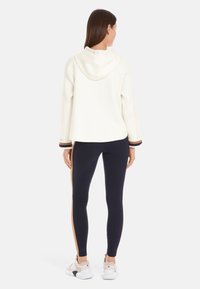 Cream hooded sweatshirt with ribbed texture and striped cuffs, paired with navy leggings featuring a gold side stripe. Both are fitted.