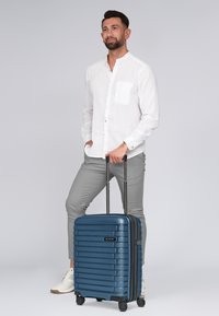 Valigia rigida di colore blu, con un design a righe texturizzate, manico retrattile e quattro ruote. Posizionata accanto a una persona in abbigliamento chiaro.