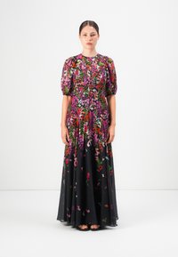 Vrouw staat in een lange, zwarte bloemenjurk met puffed korte mouwen en kleurrijke roze, rode en groene bloempatronen.