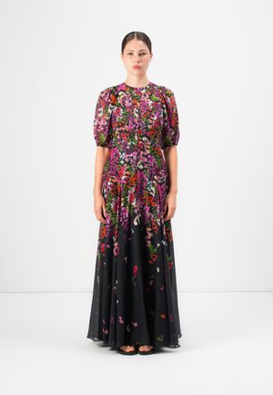 Woman standing wearing a long, black floral dress with puffed short sleeves and colorful pink, red, and green flower patterns.