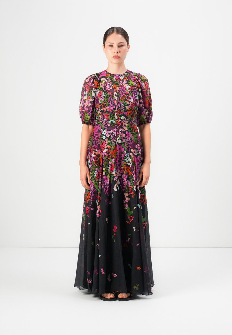 Vrouw staat in een lange, zwarte bloemenjurk met puffed korte mouwen en kleurrijke roze, rode en groene bloempatronen.